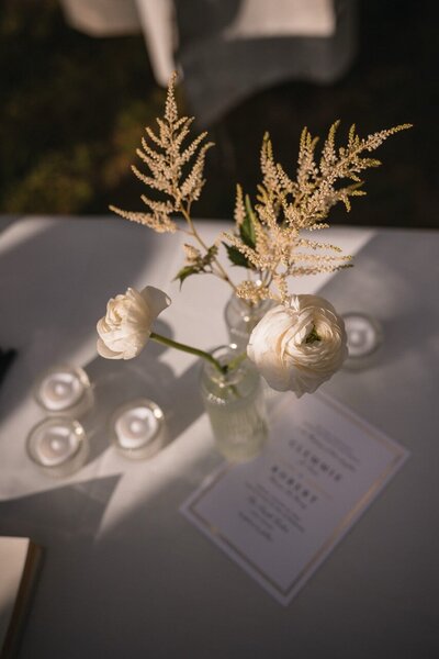 White flowers and feathery foliage in a small vase beside candles and wedding stationery.