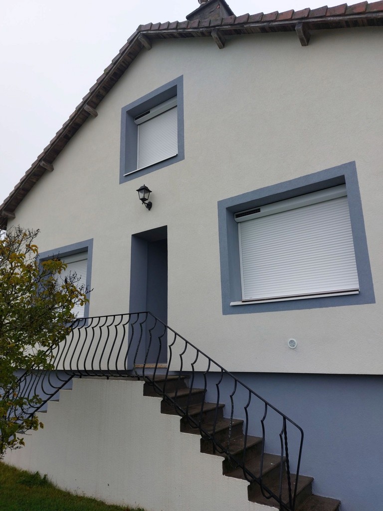 Façade d’une maison avec deux fenêtres à volets roulants fermés et une porte d’entrée encadrée de bleu