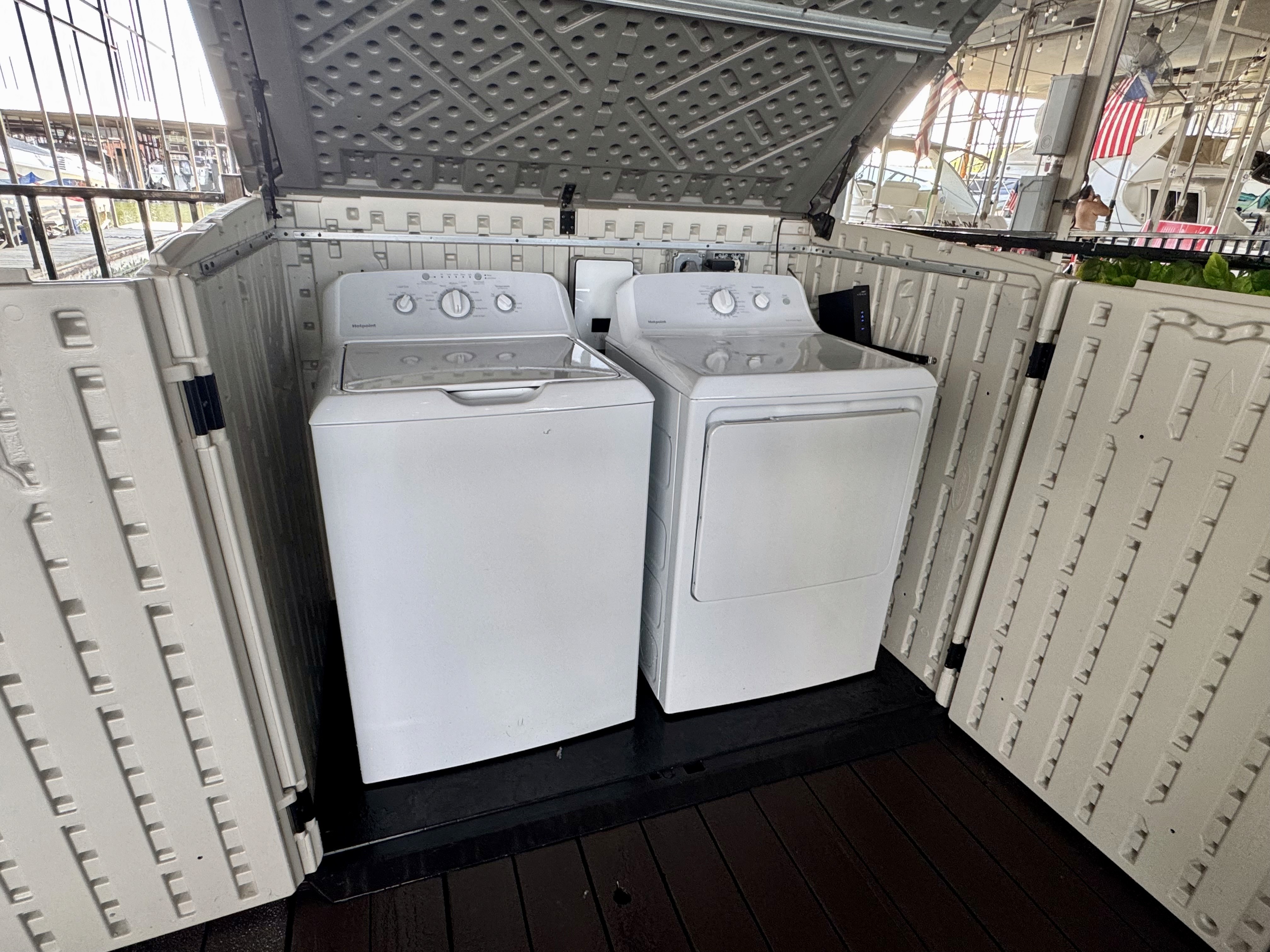 A white washing machine and dryer set housed within an open, beige outdoor storage shed on a wooden deck.