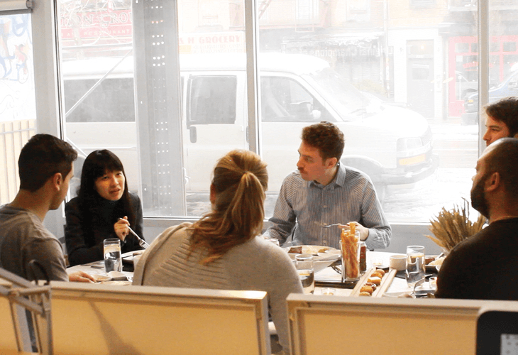 A group of six people are seated around a table in a restaurant, facing a large window that offers a view of a street scene. The table is set with plates, glasses, and small containers, suggesting a meal in progress. The individuals are dressed in casual attire. The window shows a white van parked outside and buildings across the street. The restaurant has large windows that let in natural light, and the table is surrounded by light-colored chairs.