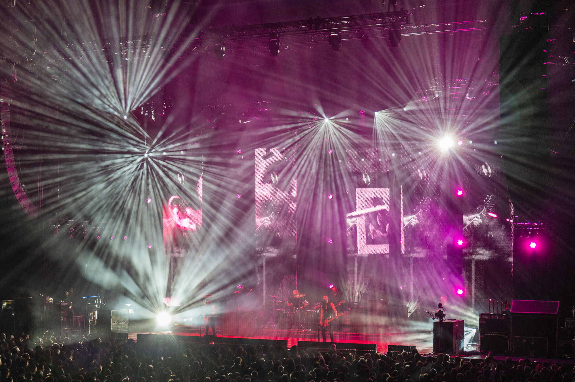 Faisceaux blancs en étoile et lumières magenta dirigés vers le public, écrans en noir et blanc, musiciens à contre-jour sur scène.