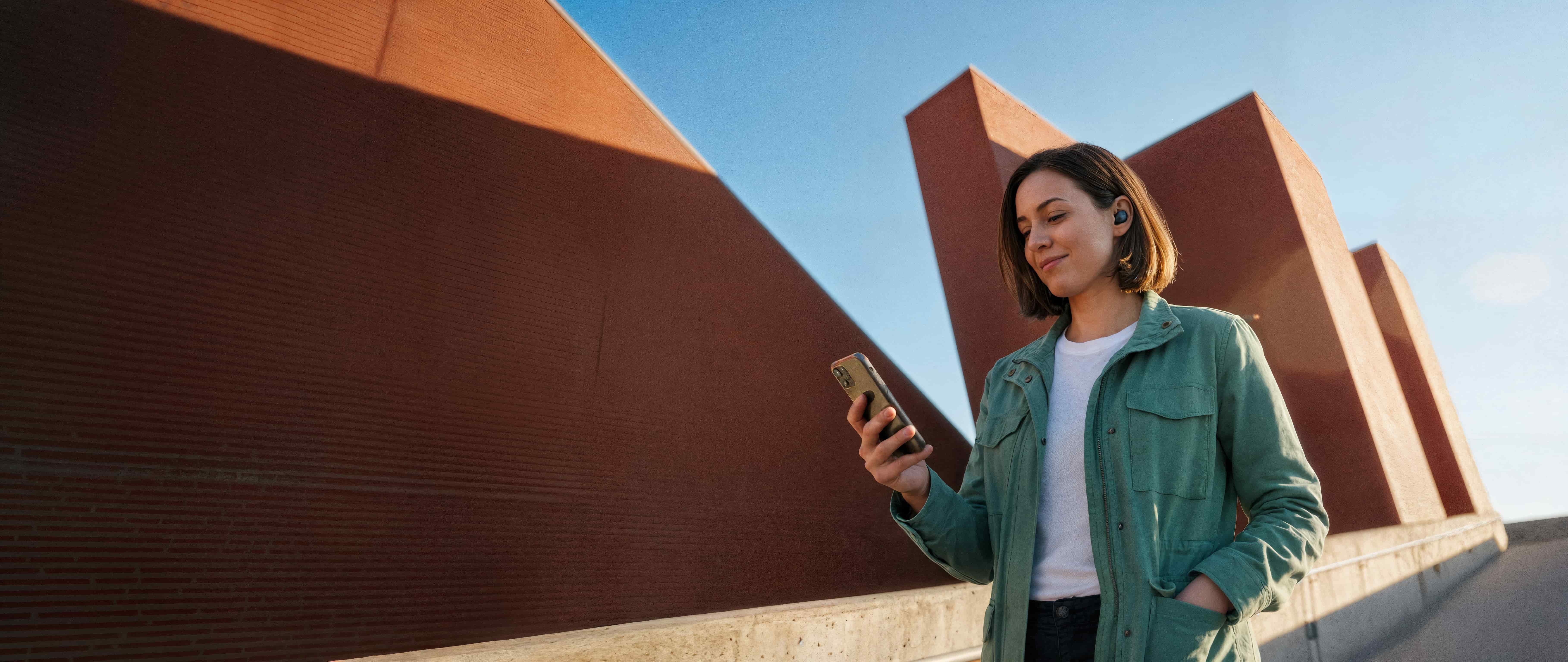 Woman using smartphone with wireless earbuds, urban background.
