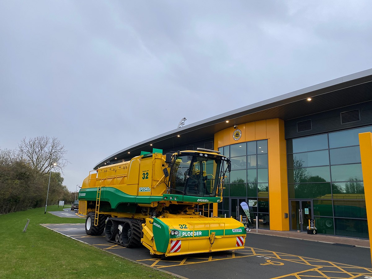A Ploeger EPD540e pea harvester (number 32) parked outside a modern commercial building with yellow and green cladding, on an overcast autumn day.