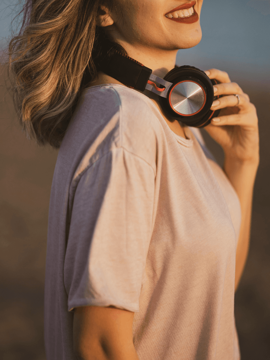 A woman wearing a light pink tshirt is standing and smiling on the beach with her headset sitting around her neck