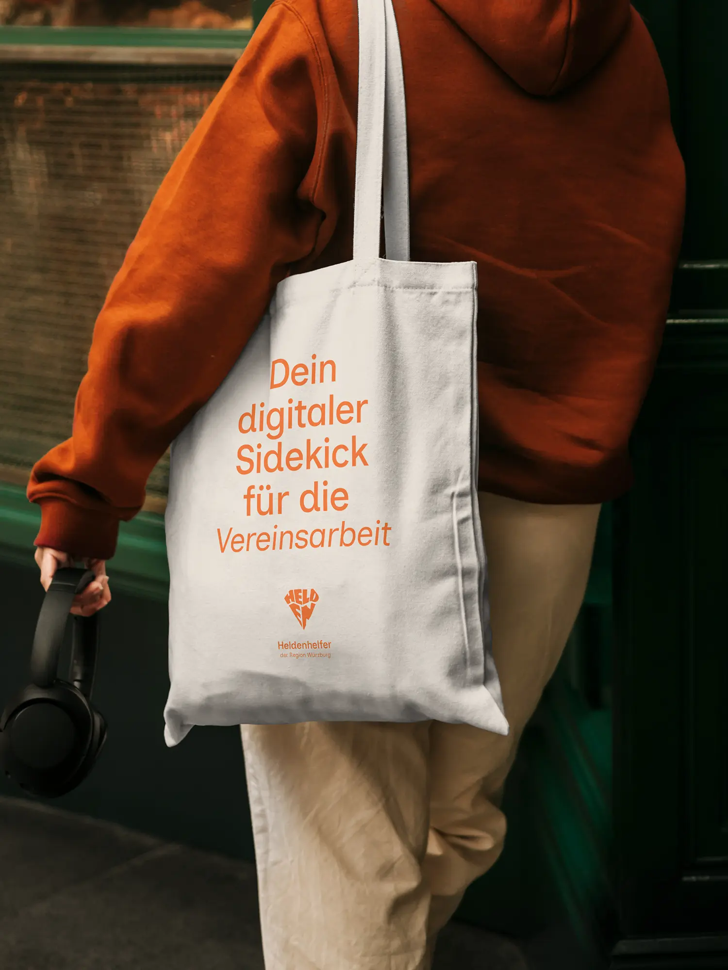 Person mit orangefarbenem Hoodie und Tragetasche „Dein digitaler Sidekick für die Vereinsarbeit“ von Heldenhelfer.