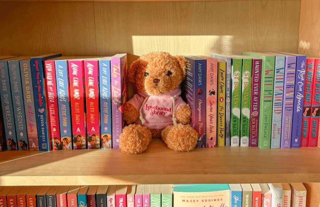 A stuffed teddy bear that is fluffy and brown with a pink hoodie on that says Lovebound Library. The bear is placed on a bookshelf between books.