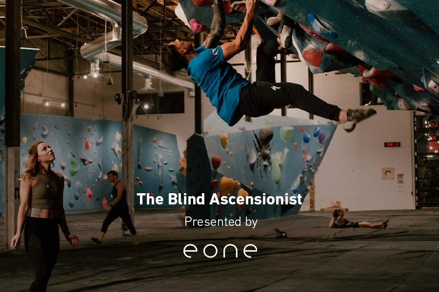 Justin Salas climbs a kilter board inside a bouldering gym. He wears a light blue t-shirt and black sports pants.