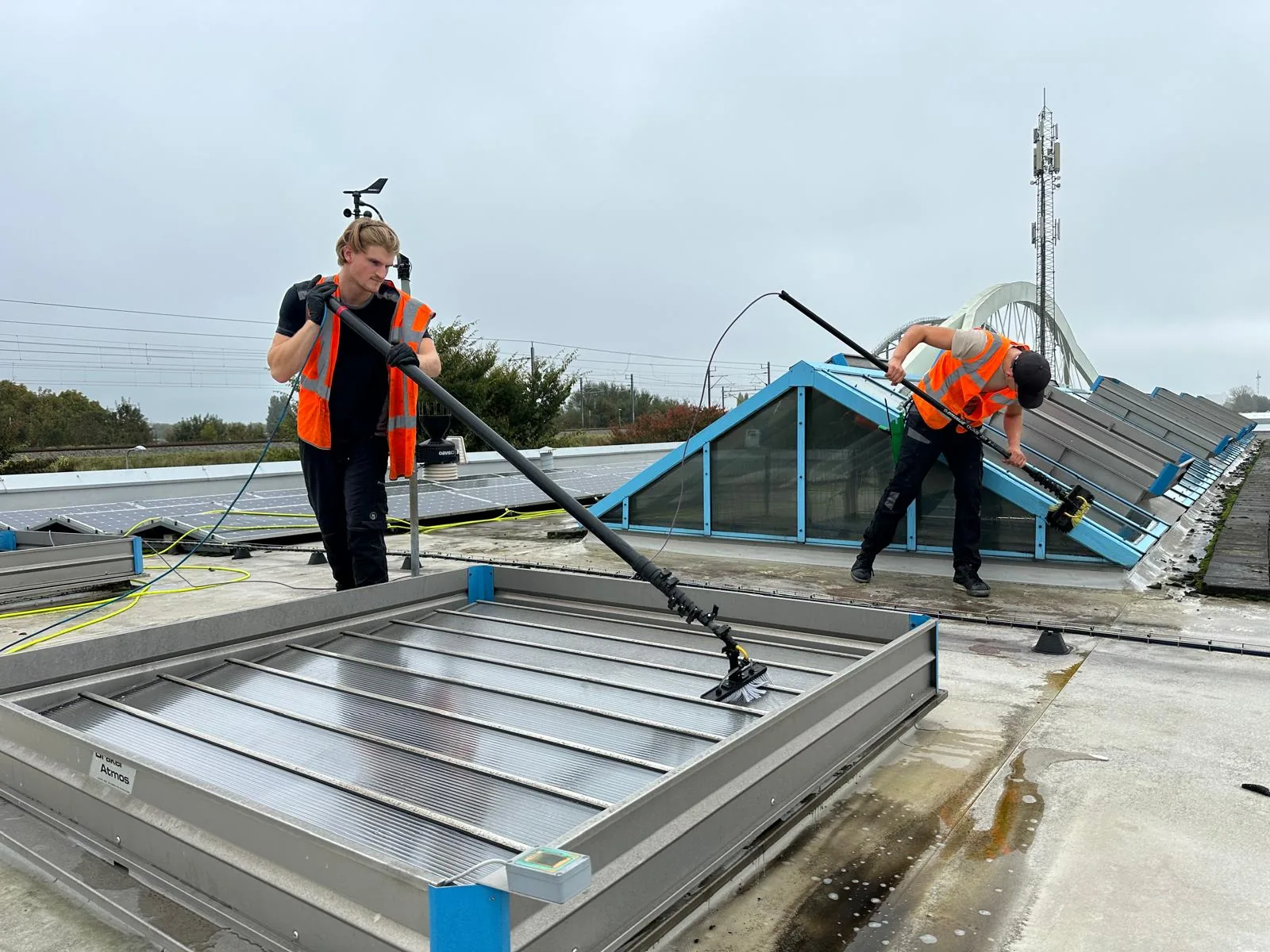 Twee medewerkers reinigen een lichtstraat op een plat dak van een bedrijfspand, gezien over het dakvlak