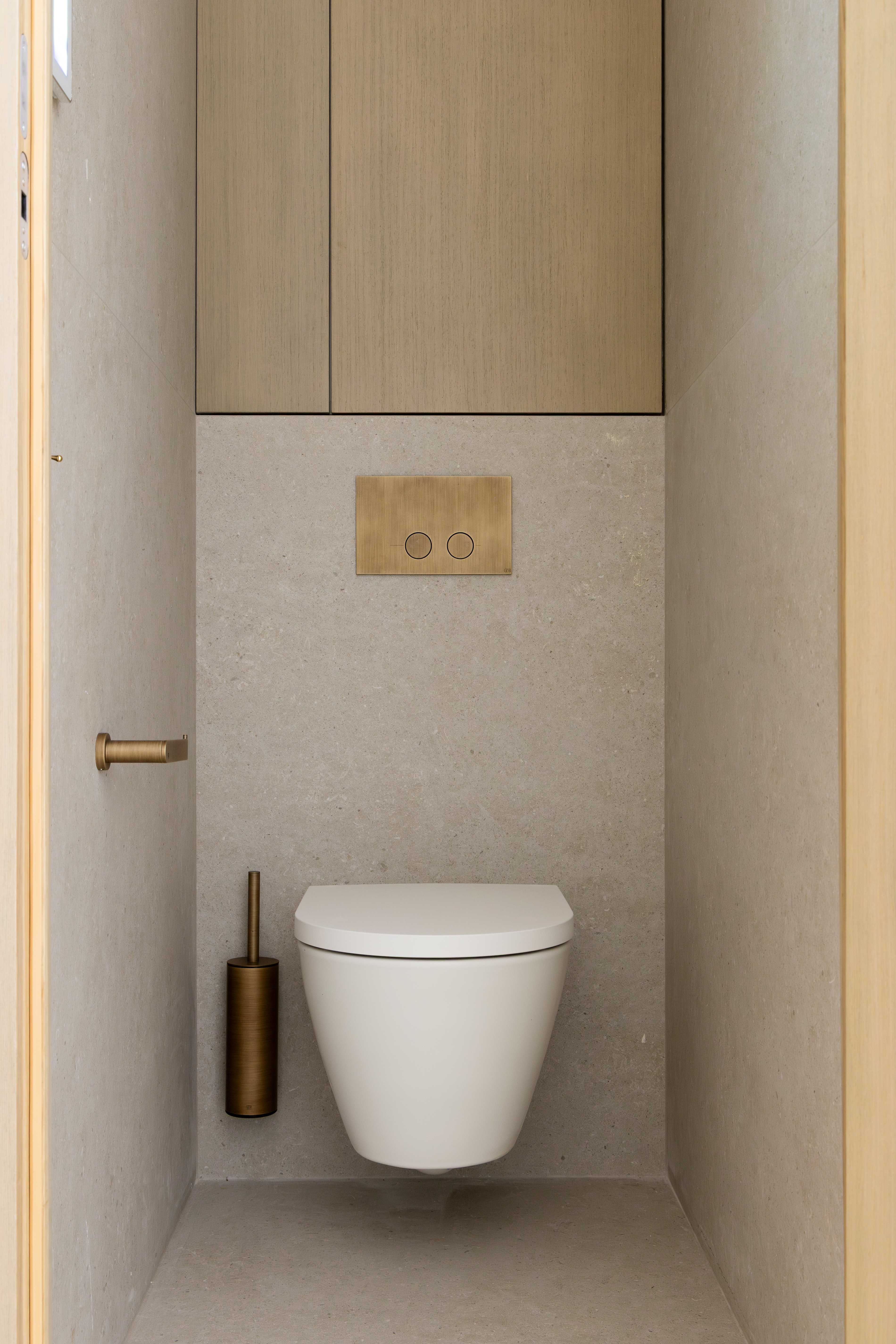 Toilettes suspendues blanches avec plaque de chasse doree, rangement en bois clair et porte-balai en laiton