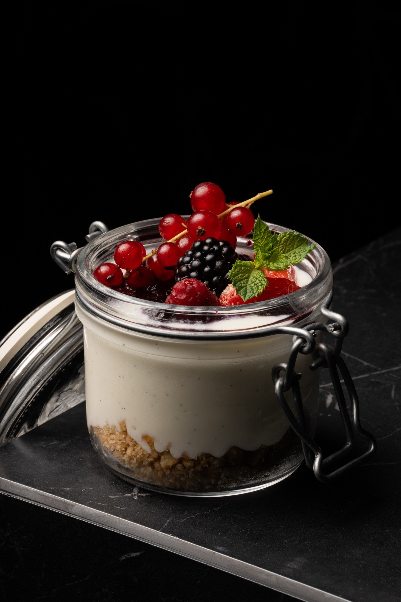 Cheesecake a strati servito in barattolo di vetro con chiusura ermetica e frutti di bosco in superficie, su tavolo grigio