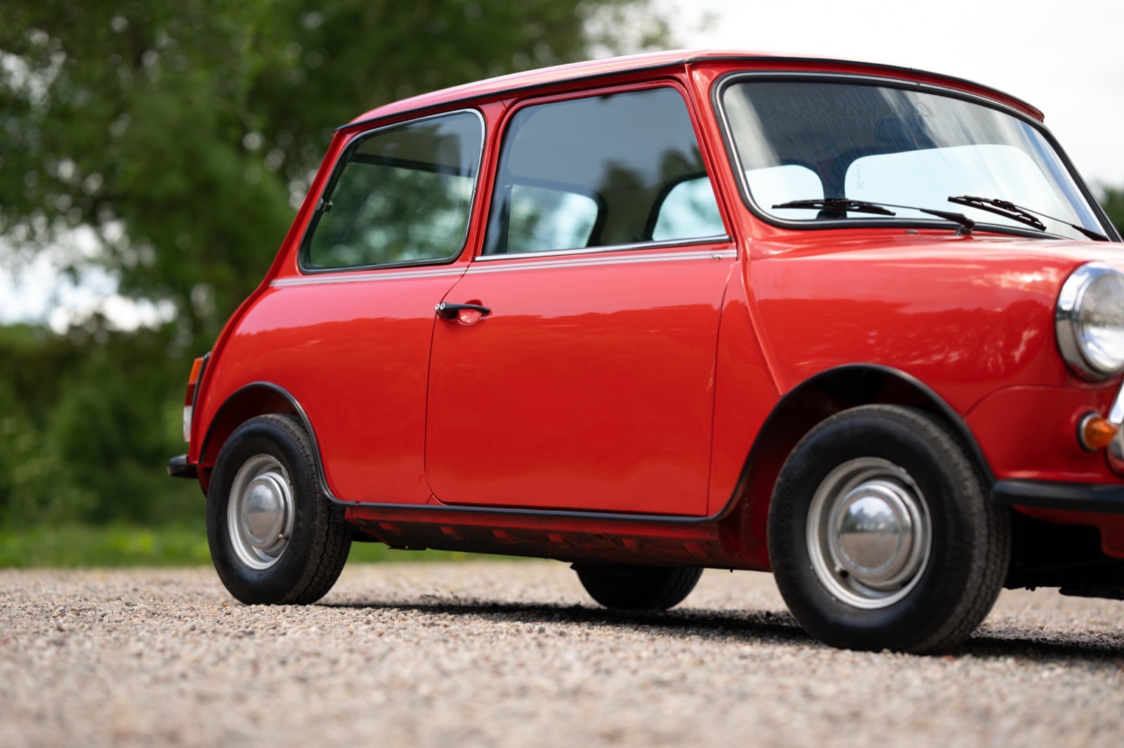 A side view of a red vintage car parked on a gravel road, with greenery in the background.