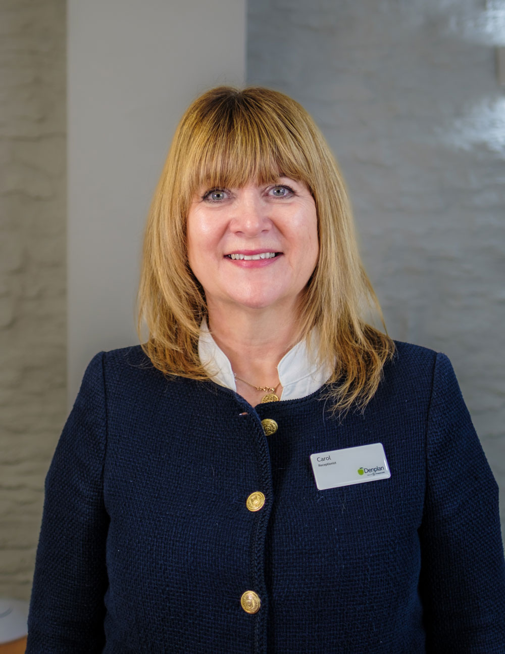 A woman with blonde hair is smiling and wearing a dark blazer with a name badge, standing in a room with a textured light-coloured wall in the background.