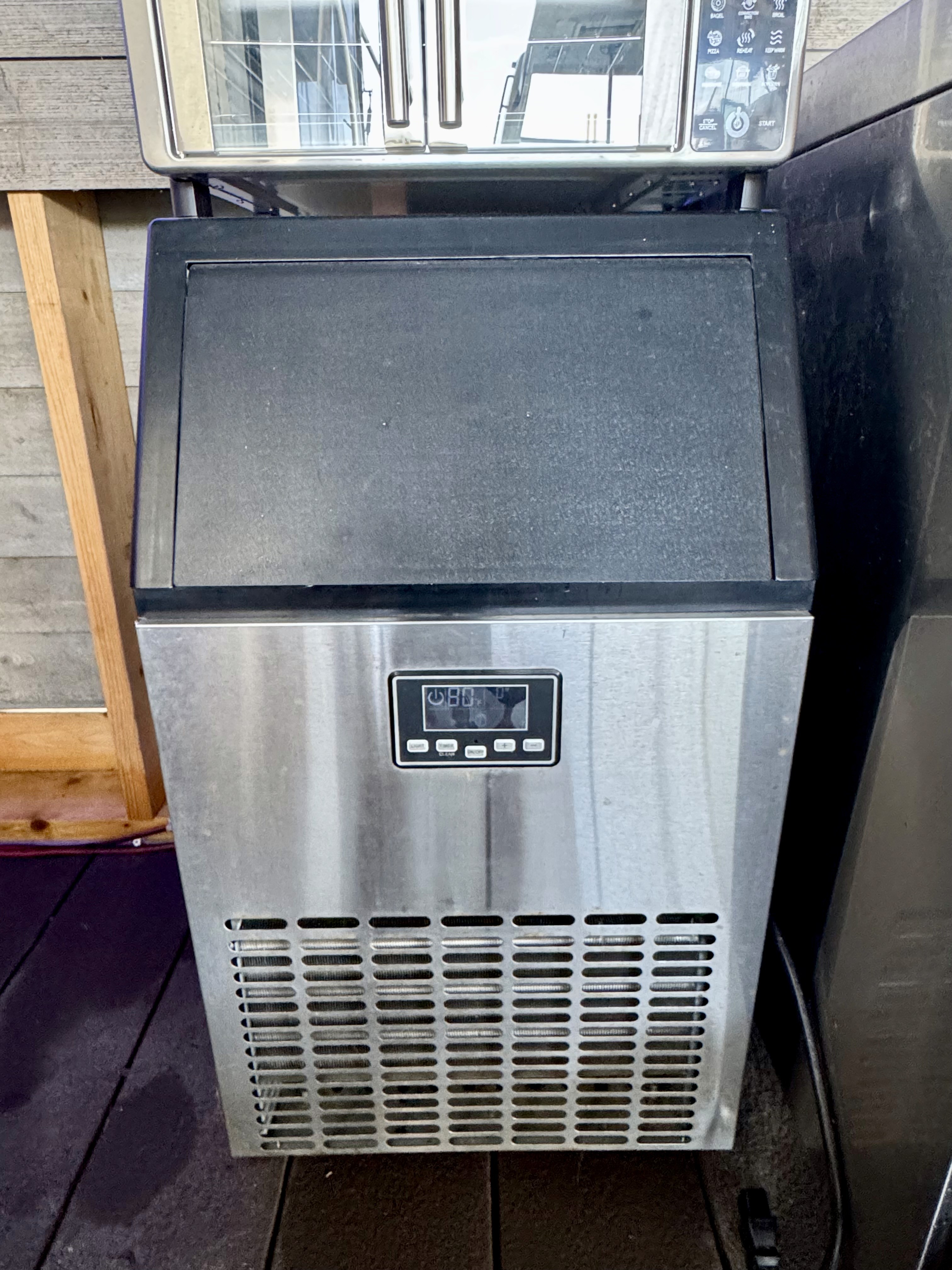 A stainless steel commercial ice maker with a digital display is situated on a wooden deck, framed by modern kitchen appliances and wooden paneling in the background.