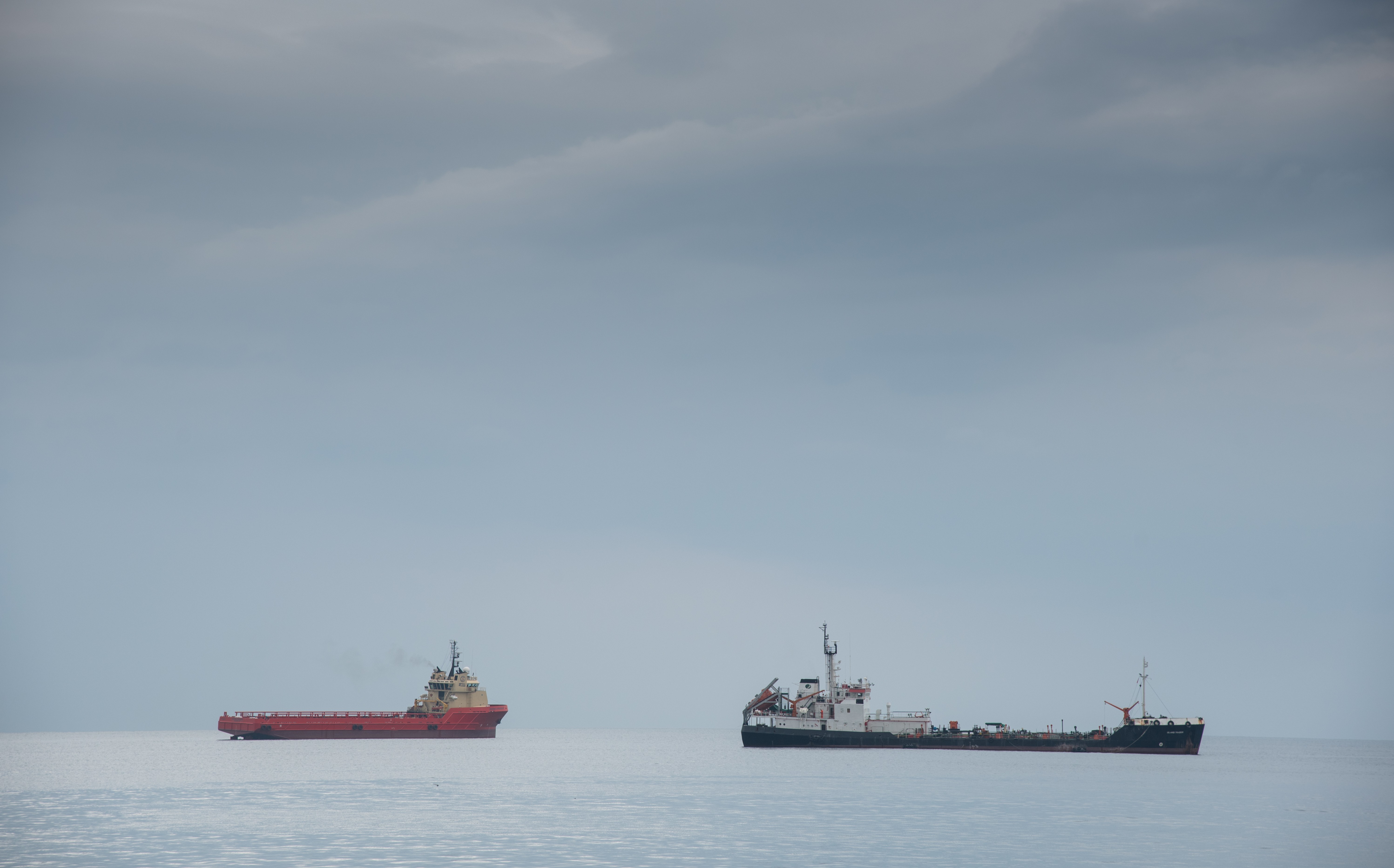 Two vessels at sea under an overcast grey sky, a red offshore supply ship on the left and a small tanker on the right