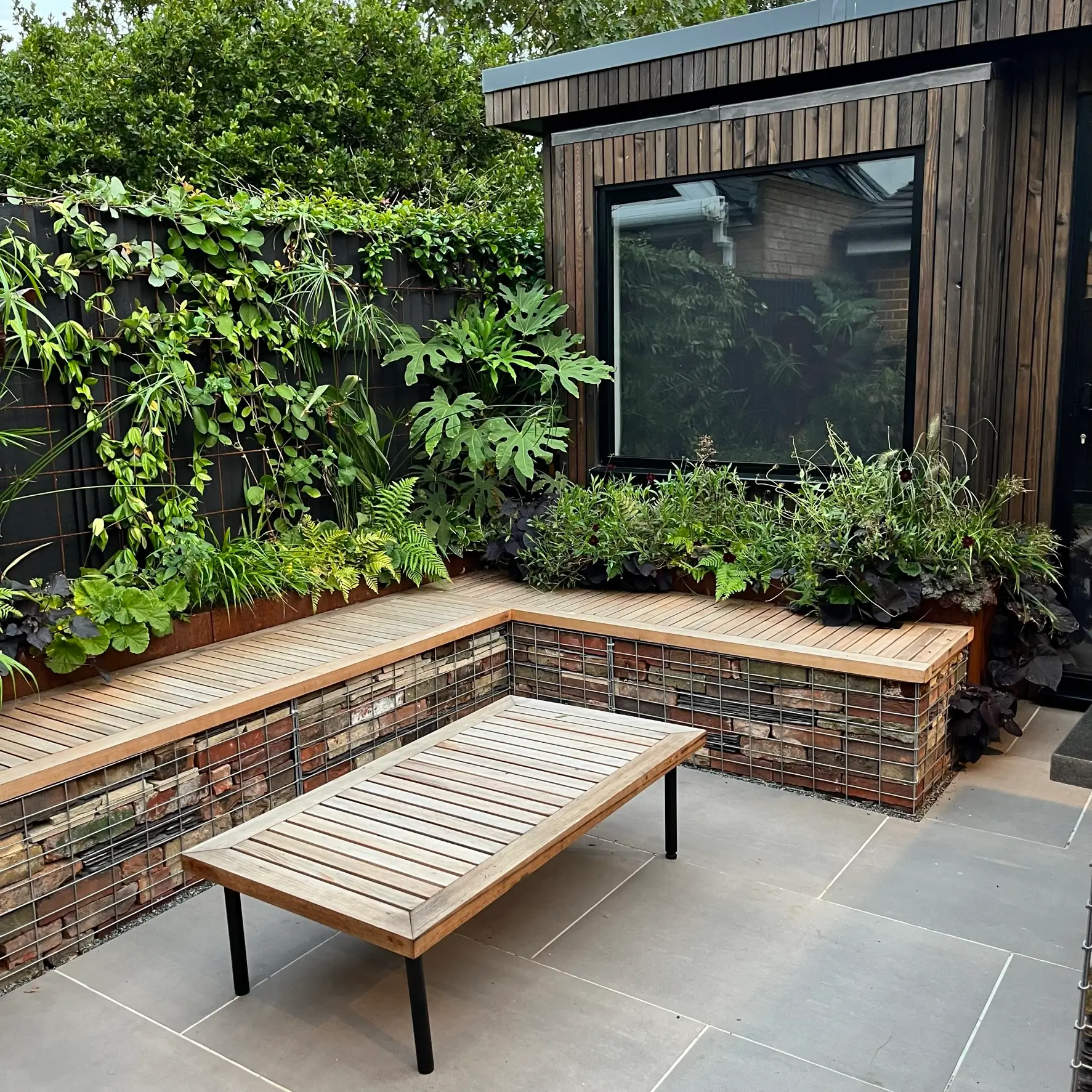 A modern outdoor space featuring a stone seating area and a wooden table surrounded by greenery and a house.