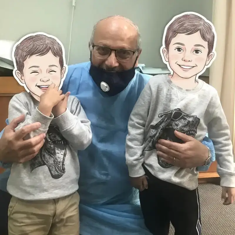 Dr Ashraf Saddik, in a medical gown and mask, embraces two young boys with illustrated faces and matching grey sweaters, at his family medical clinic in Coburg, serving Melbourne's northern suburbs.