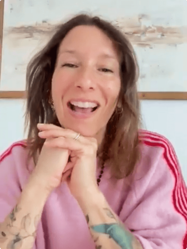 Une femme souriante en sweat-shirt rose à rayures rouges, les mains jointes sous le menton, avec des tatouages.