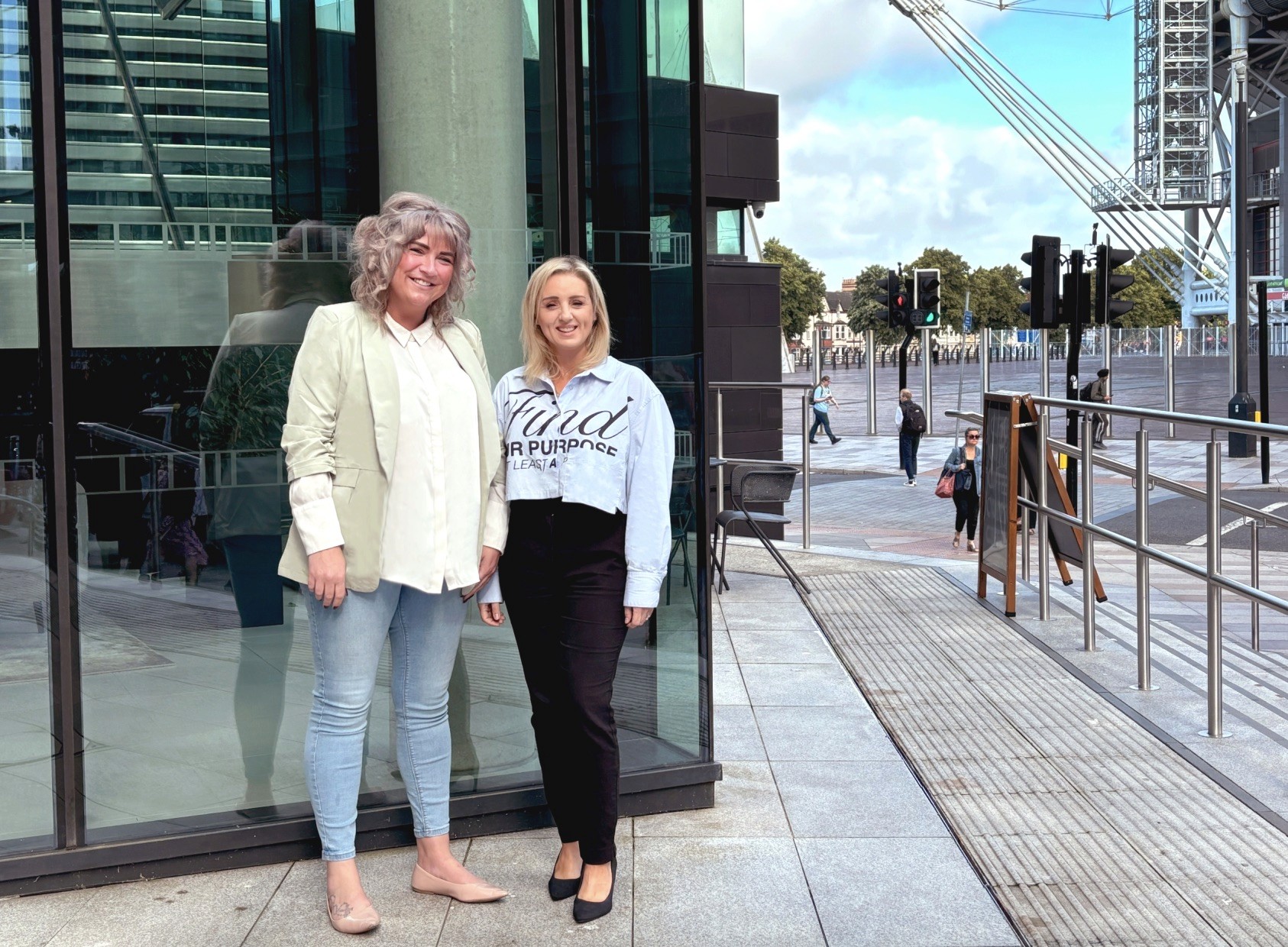 Two people standing near a glass building with another person walking.