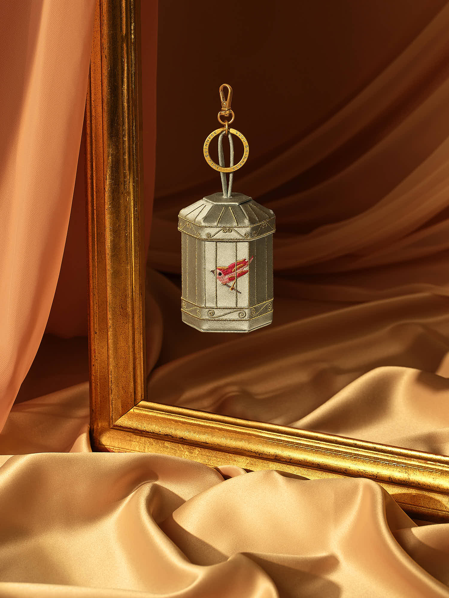 Luxury birdcage keychain with gold frame and silky orange drapery background.