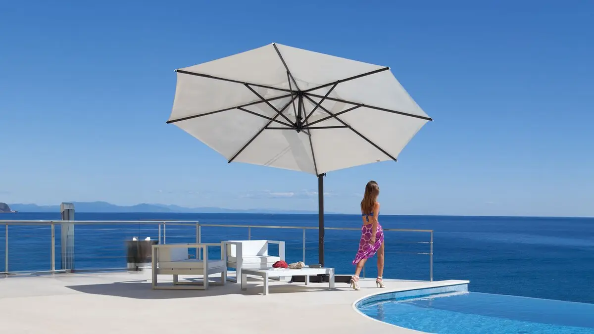 Mujer con vestido veraniego camina al borde de una piscina infinita que mira al mar. Tumbona y parasol Flexy de Fim Umbrellas
