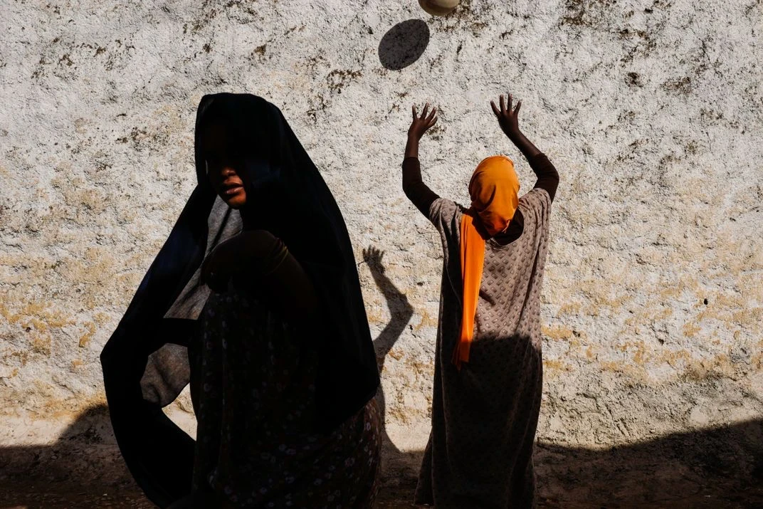Two women in headscarves stand against a textured white wall, one draped in dark fabric, the other in orange with arms raised toward a dark disc suspended mid-air above her