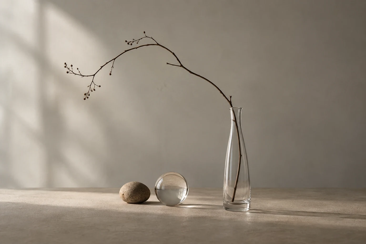 Single long curved twig in a clear narrow glass vase beside a small rounded stone and a clear glass sphere, warm gray tabletop