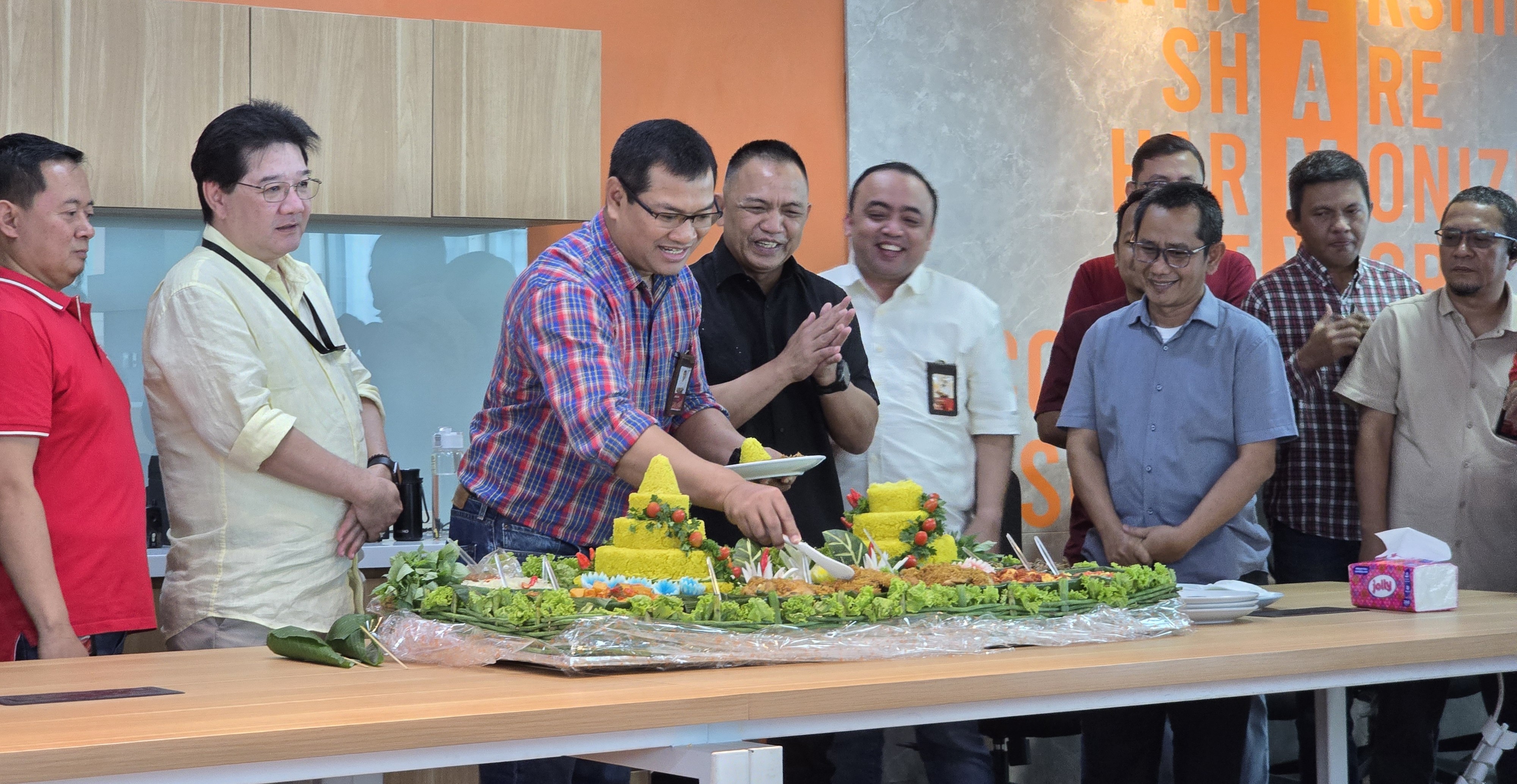 Sejumlah karyawan berkumpul di ruang kantor modern saat acara syukuran, dengan seorang pria memotong tumpeng nasi kuning di atas meja panjang, dikelilingi rekan-rekan yang tersenyum dan bertepuk tangan