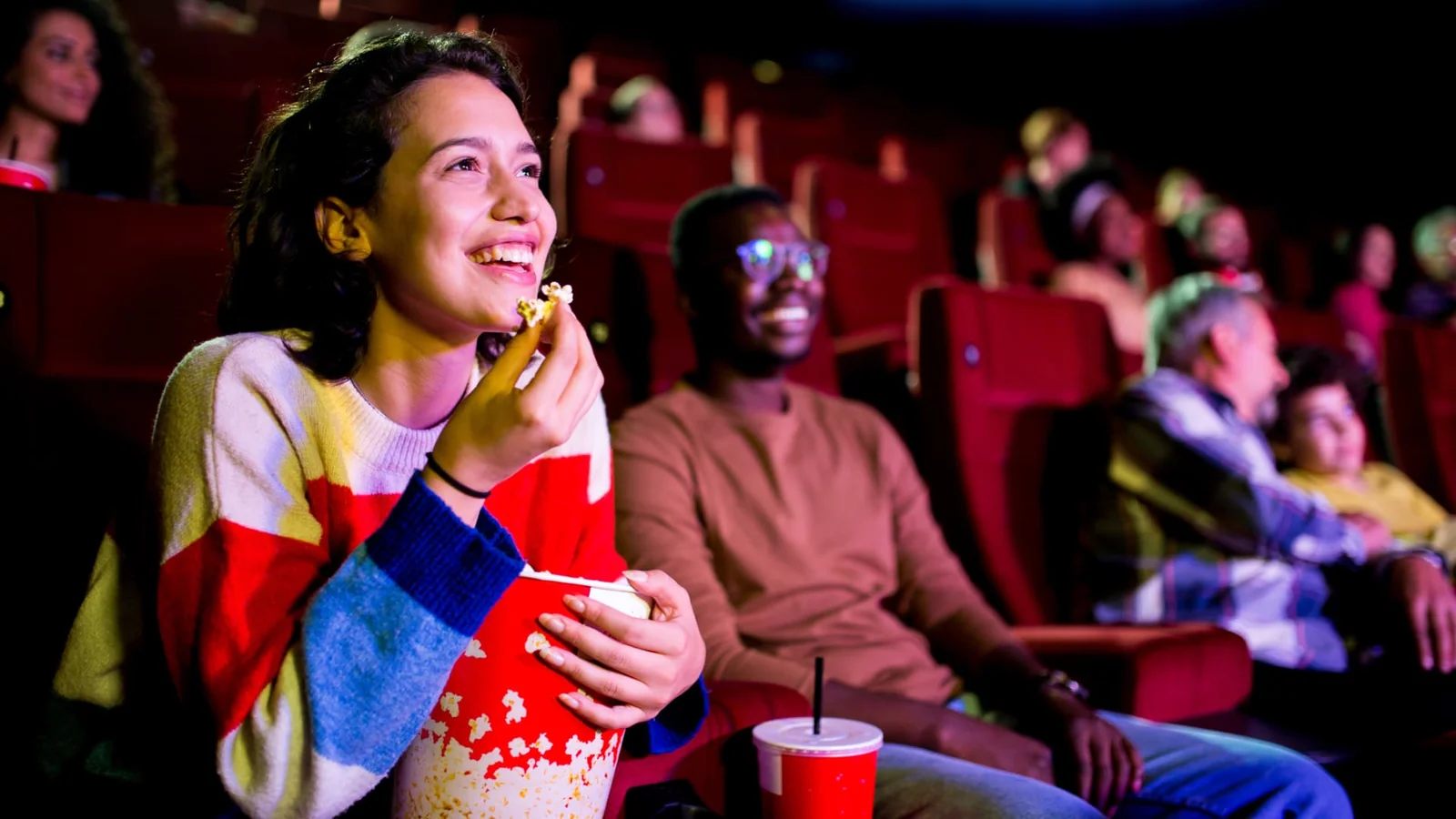 Young woman enjoying popcorn and movie in theater with other audience members seated in red chairs.