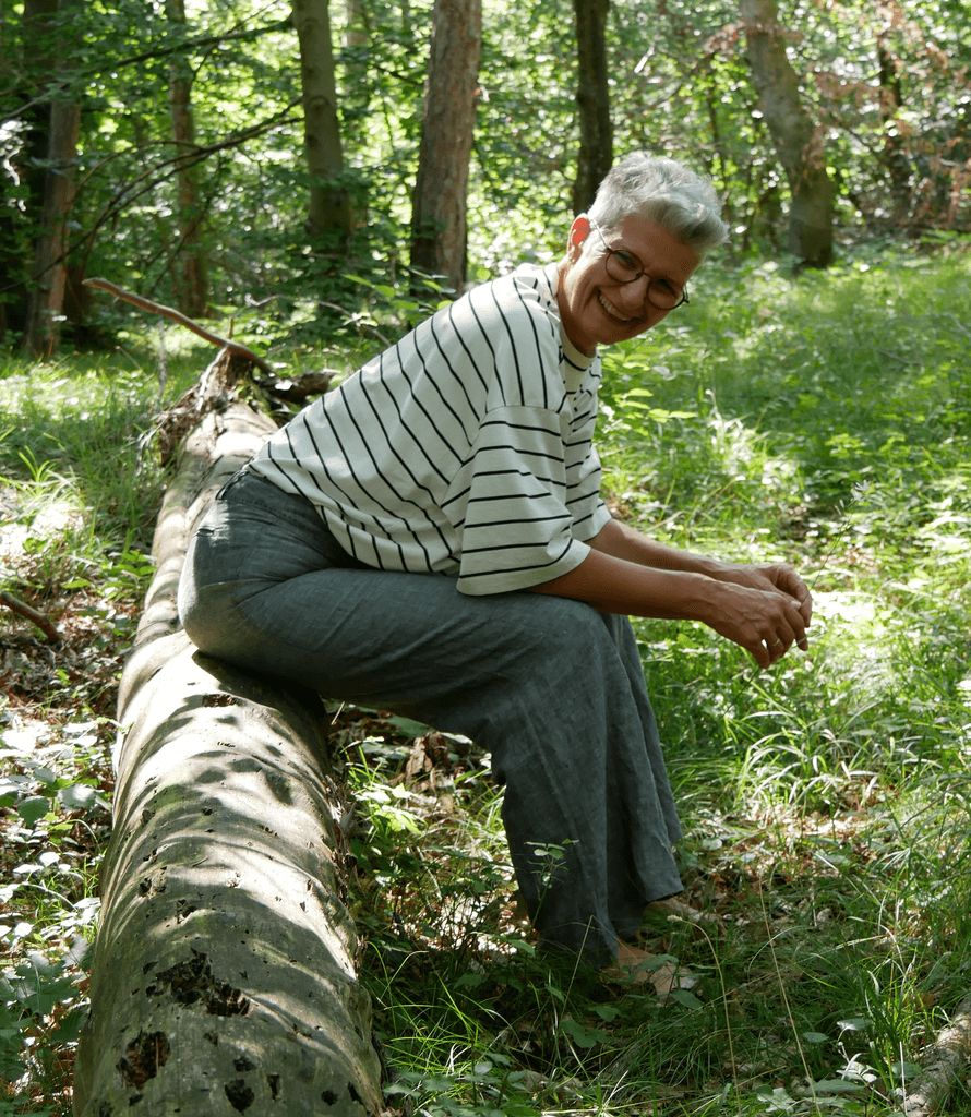 Person mit grauen Haaren und Brille sitzt lächelnd auf einem umgestürzten Baumstamm im Wald. Sie trägt ein pinkfarbenes Oberteil und eine graue Hose, umgeben von dichtem grünen Laub.