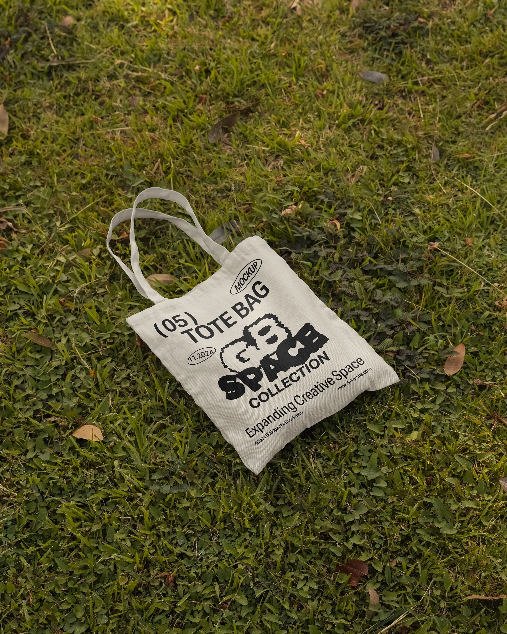 A white tote bag with bold black graphics and text, lying on green grass with scattered leaves. The natural outdoor setting highlights the eco-friendly and casual vibe of the bag.