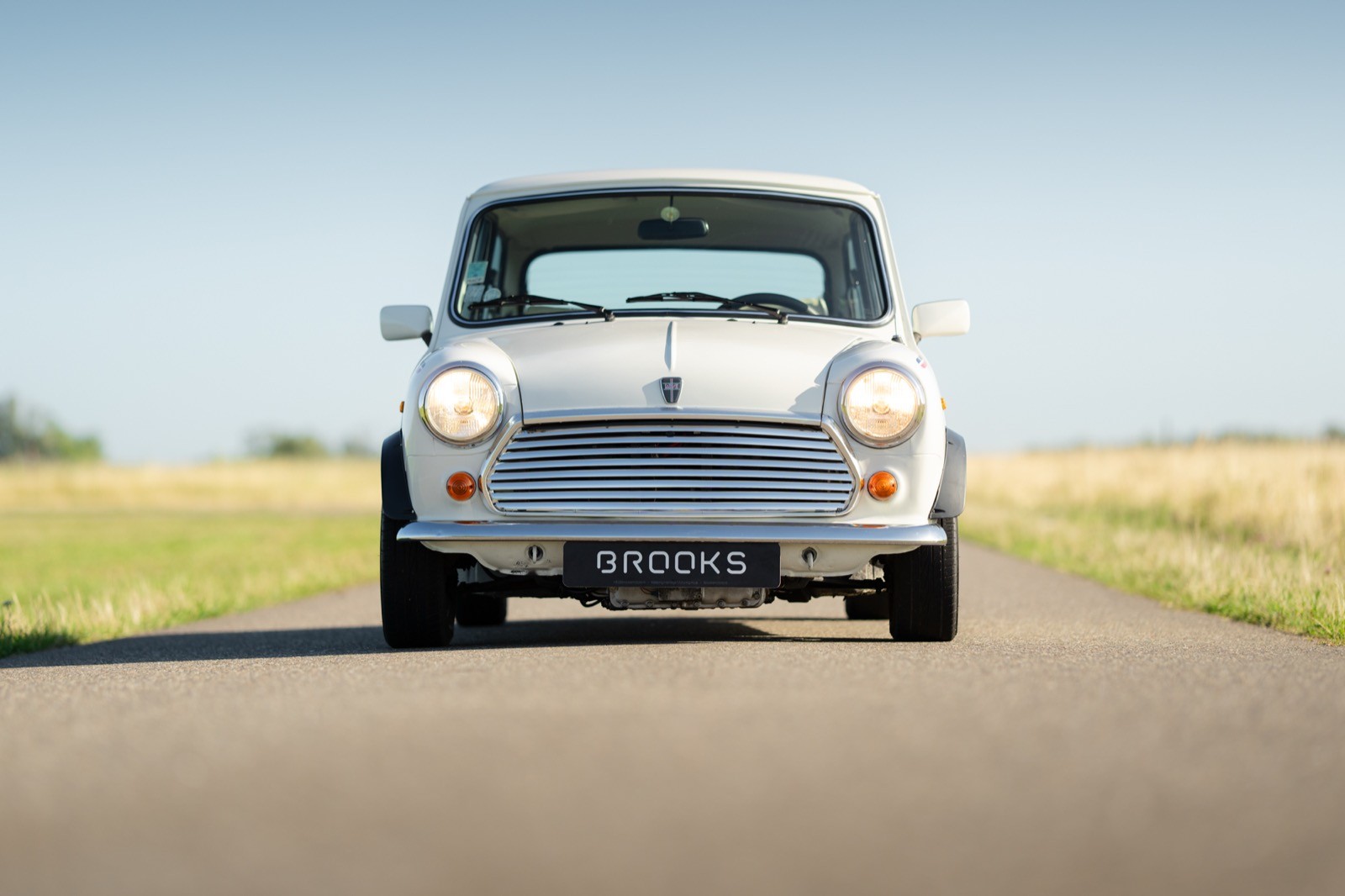 A classic car drives down a smooth road, surrounded by open fields and a clear blue sky.