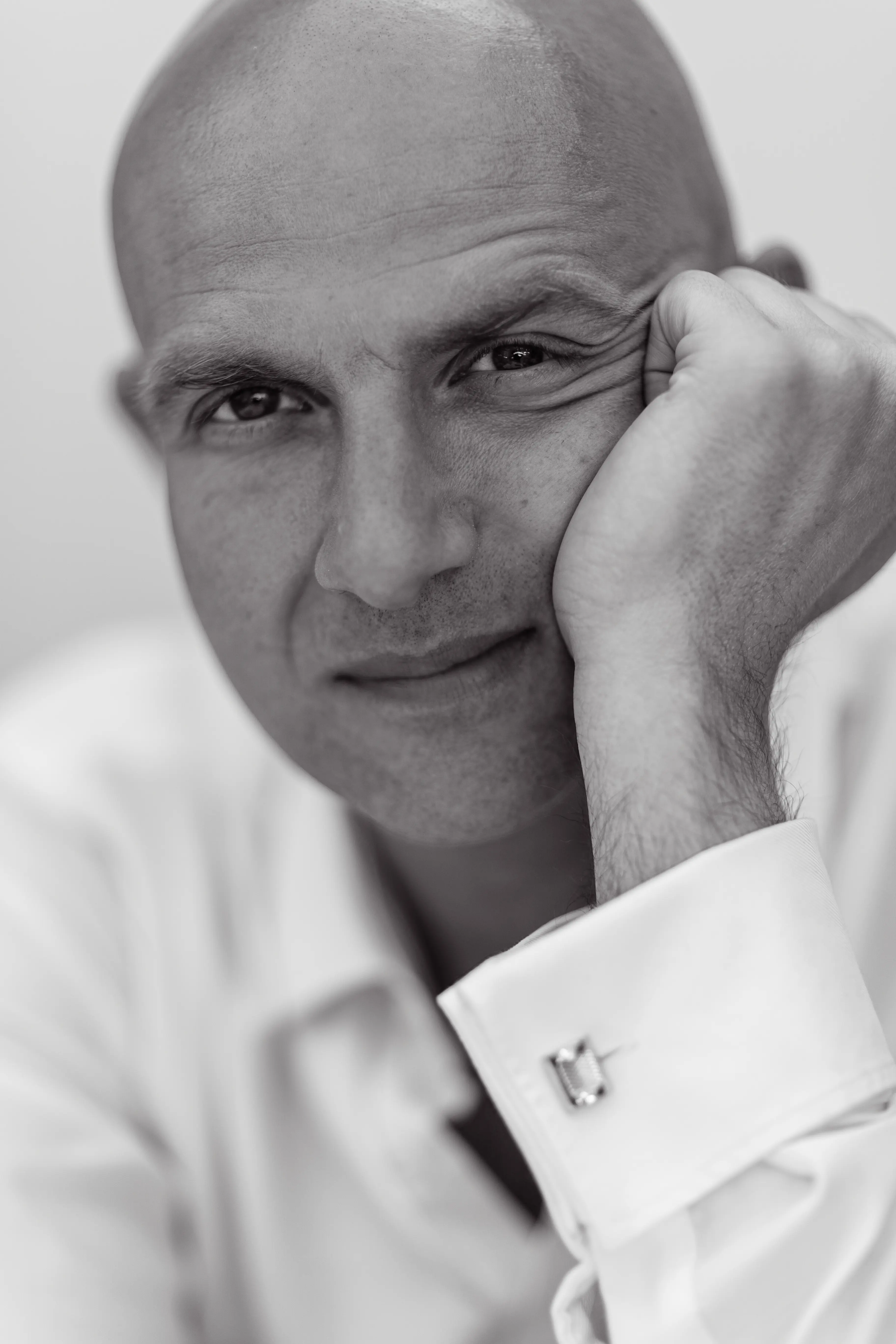 Czarno-biały portret w zbliżeniu łysego mężczyzny w białej koszuli, opierającego głowę na dłoni i patrzącego bezpośrednio w kamerę z lekkim, zamyślonym wyrazem twarzy.