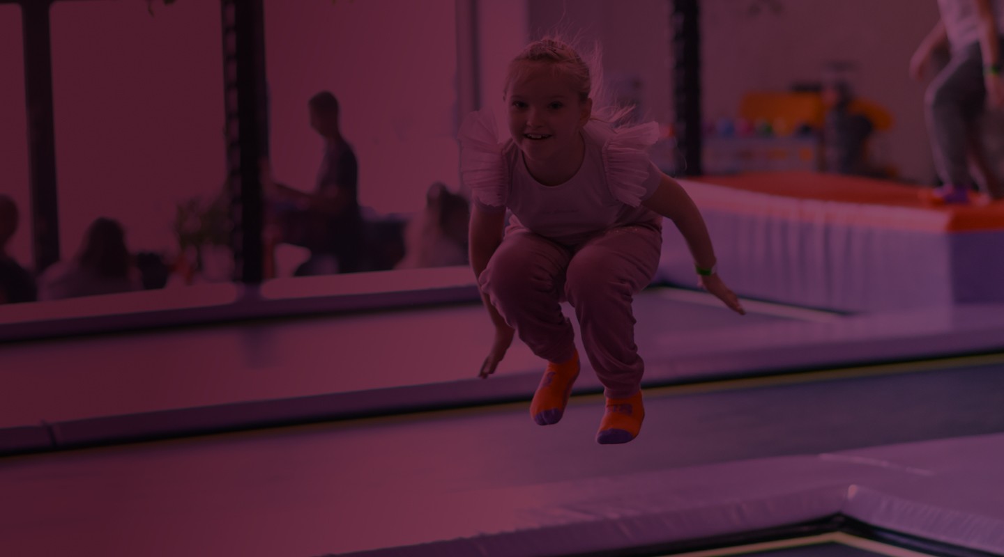girl smiling jumping on trempolines wearing pink tishirt, grey pants and orange ABS socks