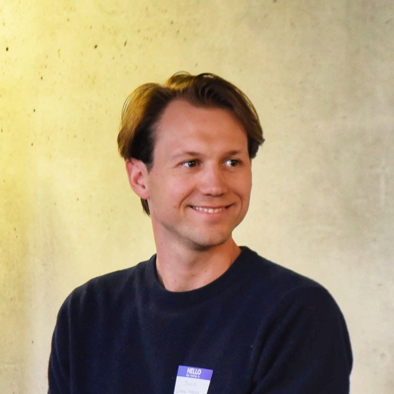 Smiling man wearing glasses and a grey shirt standing with folded arms against a neutral background.