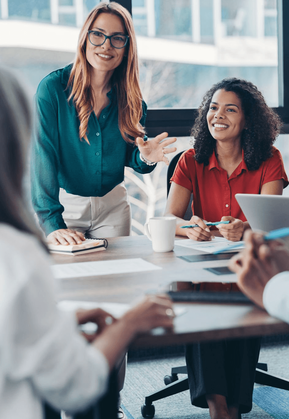 A man wearing glasses and a blazer working at a desk, writing in a notebook beside a computer monitor in a modern office.