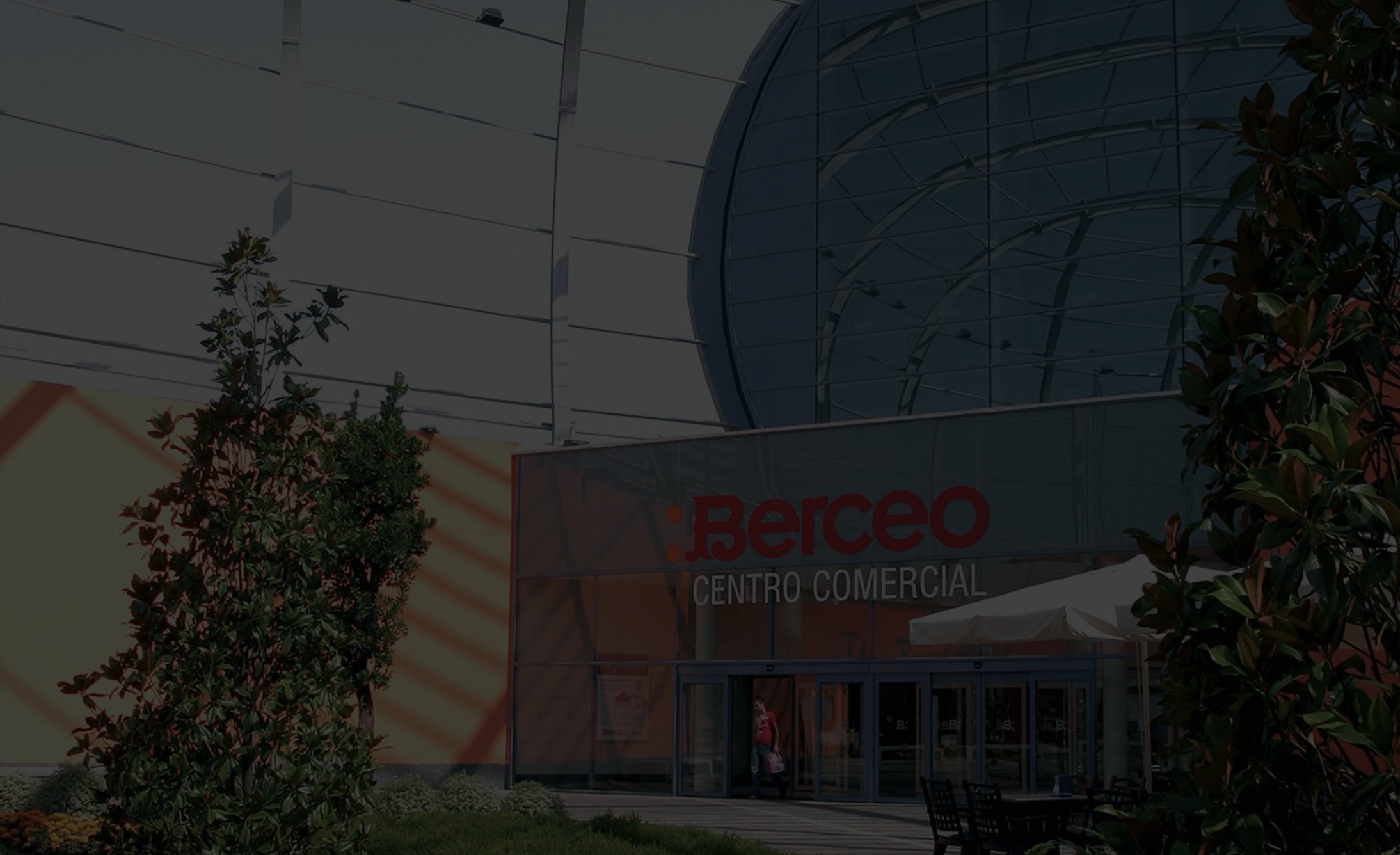 Exterior of a shopping center with "Berceo Centro Comercial" on the facade. Modern glass architecture, plants in the foreground, sunny day.