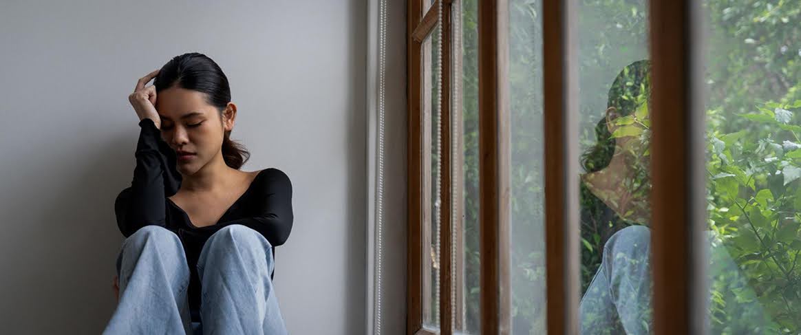 A person sits with a reflective expression near a window overlooking greenery, suggesting themes of stress and contemplation.