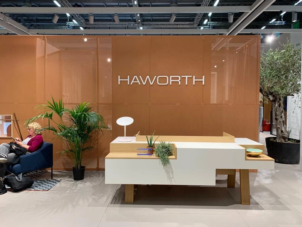 Reception of the HAWORTH ORGATEC stand with white lettering on copper mesh walls, a white counter with plants and an olive tree.