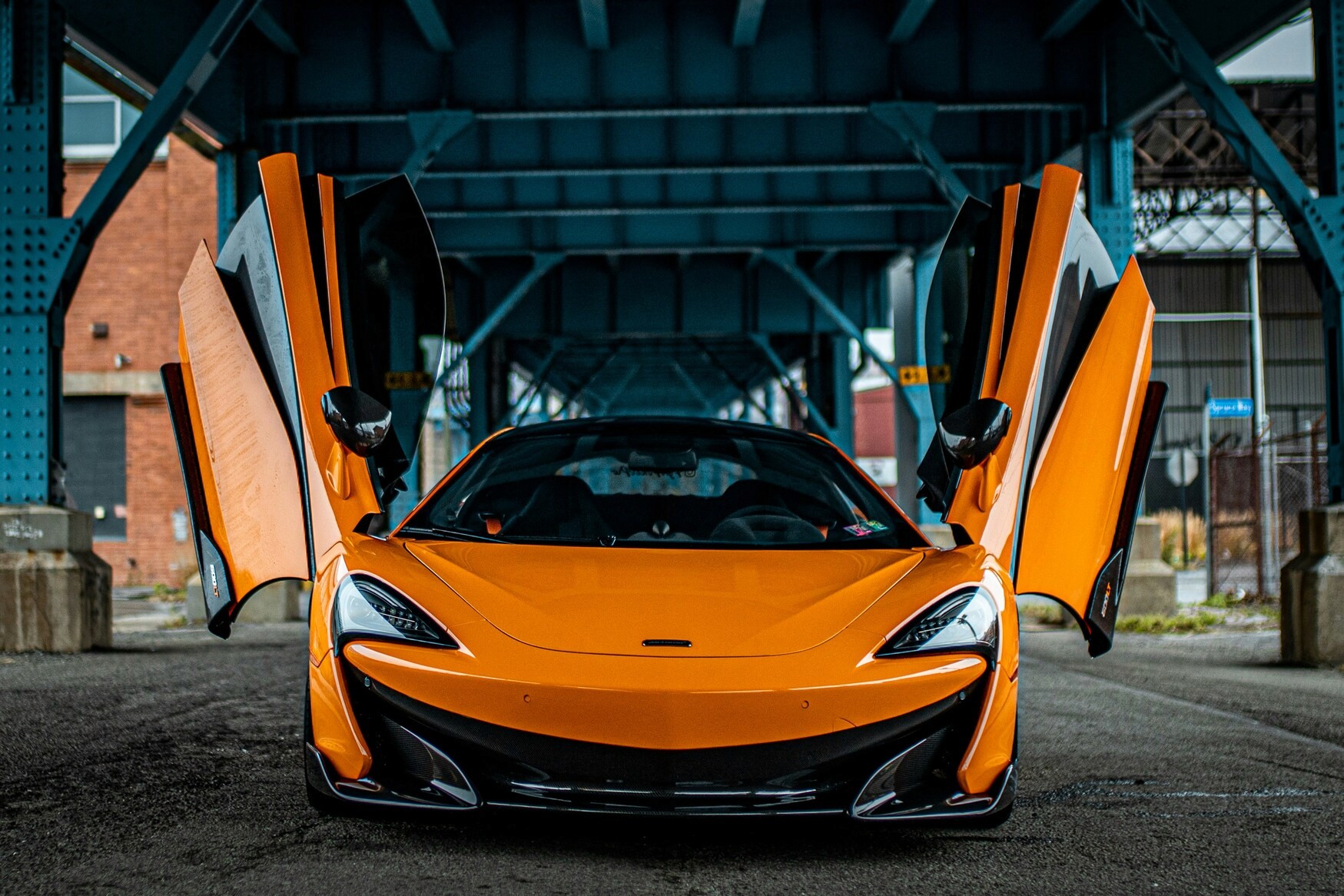 A sleek, orange sports car with gull-wing doors is parked under a steel bridge, showcasing its aerodynamic design and vibrant color.