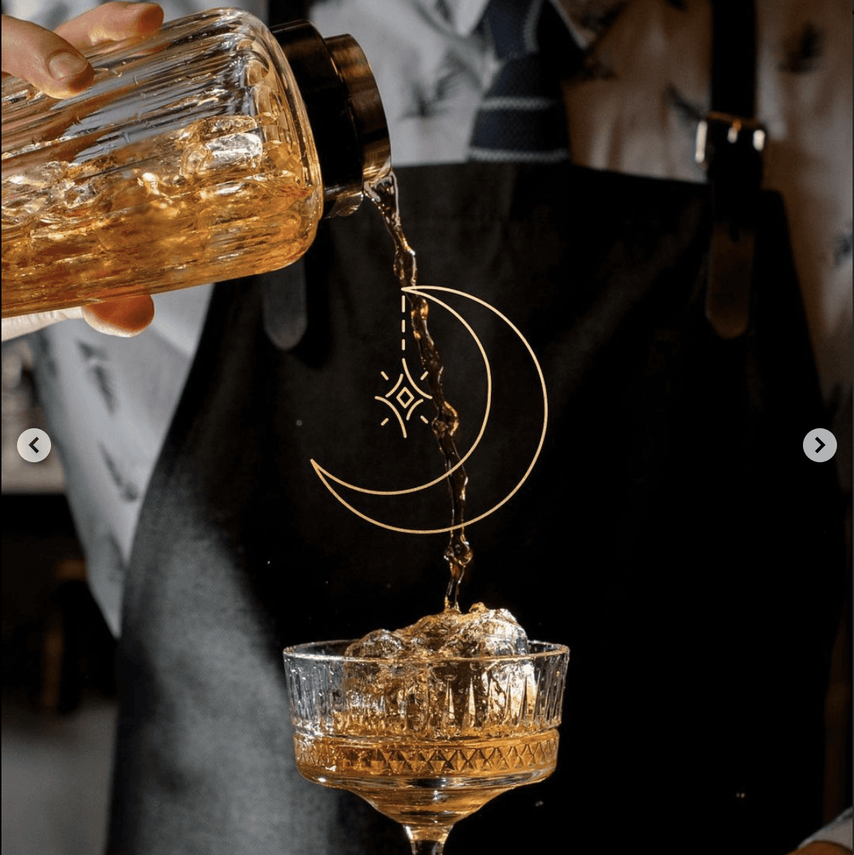 A person in an apron pouring a golden liquid from a glass container into a decorative cup.