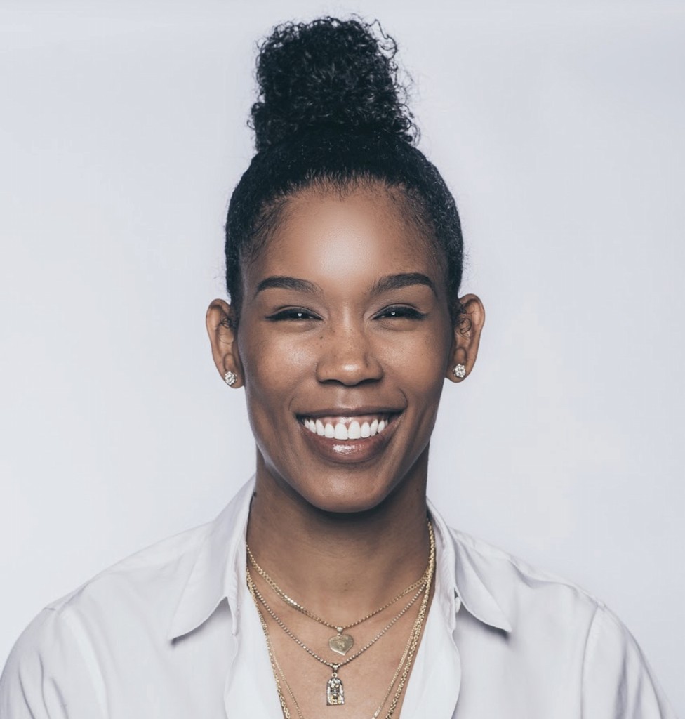 Ty Young with curly hair in a bun, wearing a white shirt and multiple gold necklaces, smiles at the camera against a plain light background.