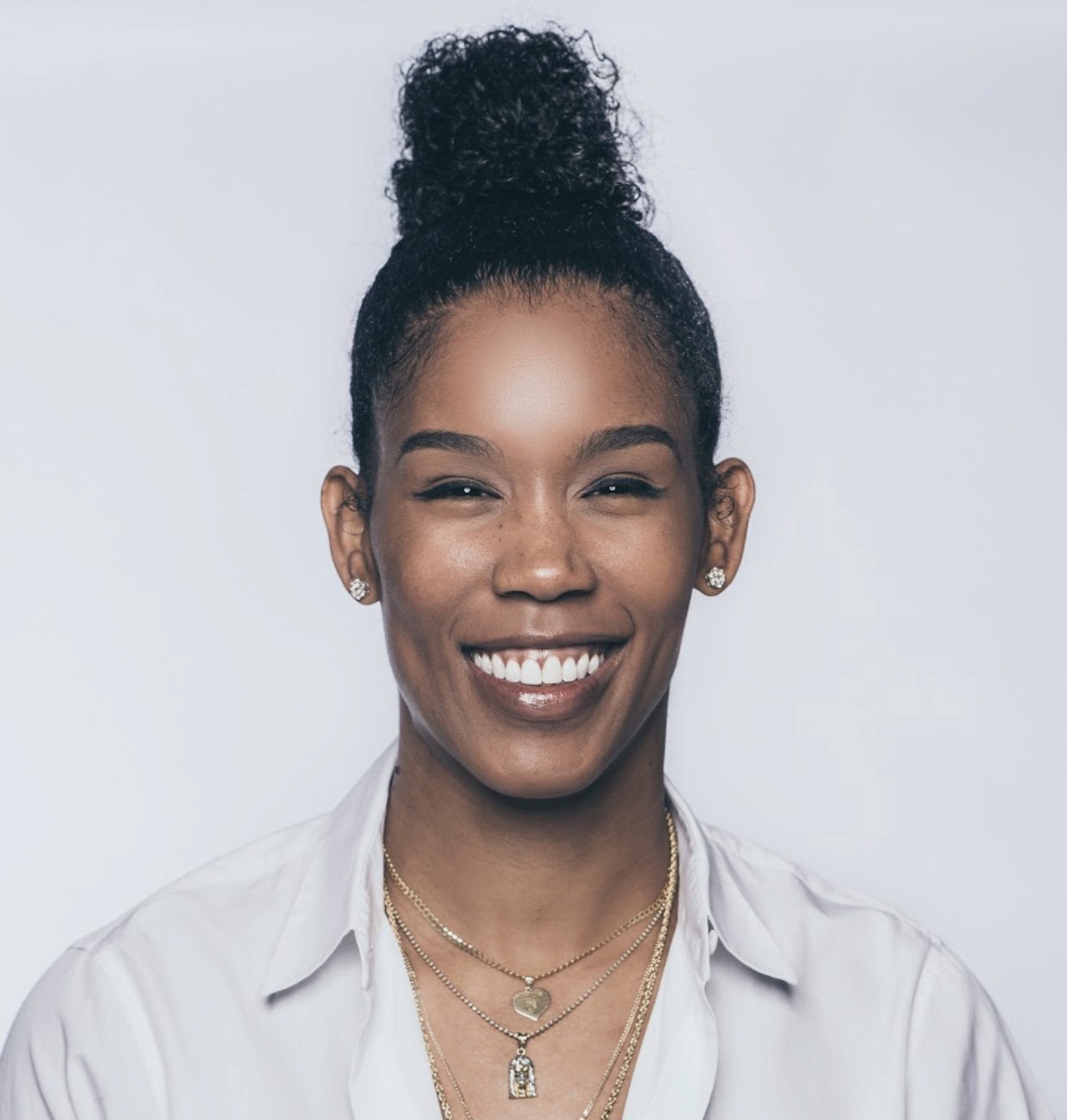 Ty Young with curly hair in a bun, wearing a white shirt and multiple gold necklaces, smiles at the camera against a plain light background.