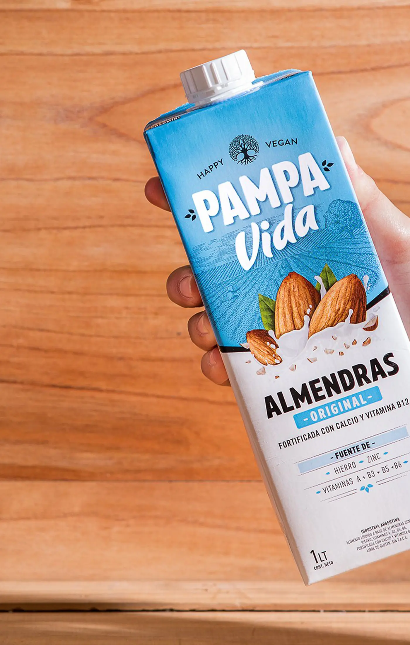 A hand holds a Pampa Vida almond milk carton against a wooden background. The packaging is blue and white, featuring almonds and text about nutrients.