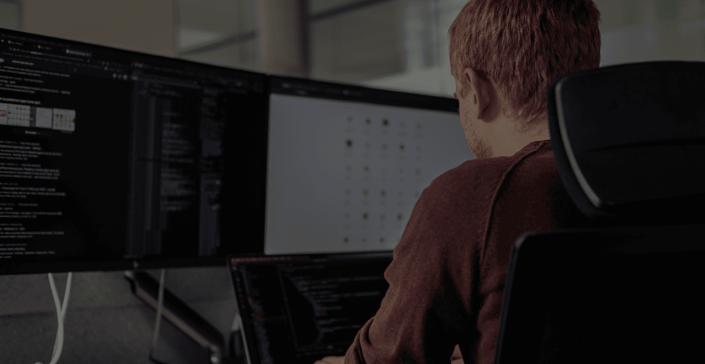 Person sitzt vor einem Laptop und zwei Bildschirmen und coded