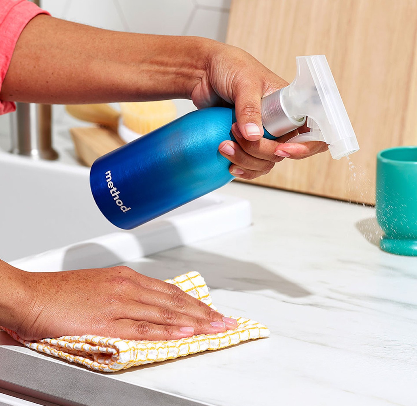 Hand spraying a blue aluminum spray bottle over a marble counter while the other hand wipes with a cloth