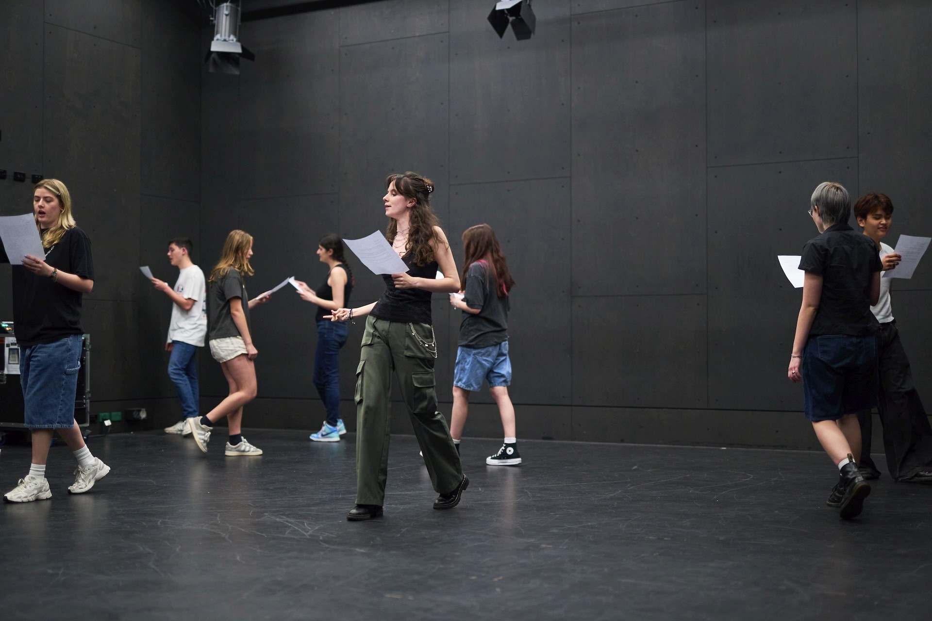 A group of teenagers walk through a rehearsal exercise while reading scripts in a large studio space.