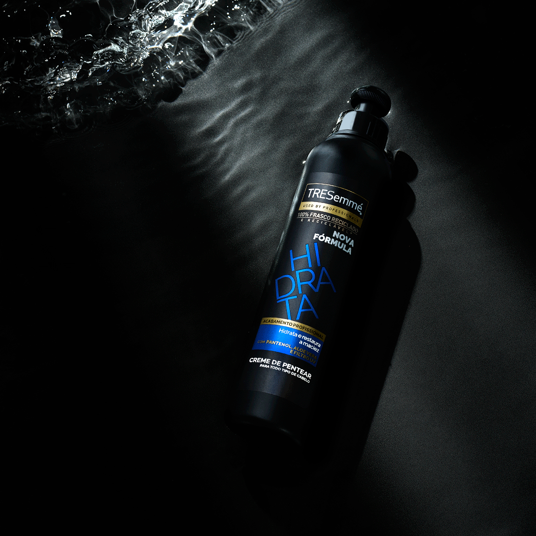 A dark shampoo bottle floating in puddles of waved water partially illuminated, with a textured surface beneath.