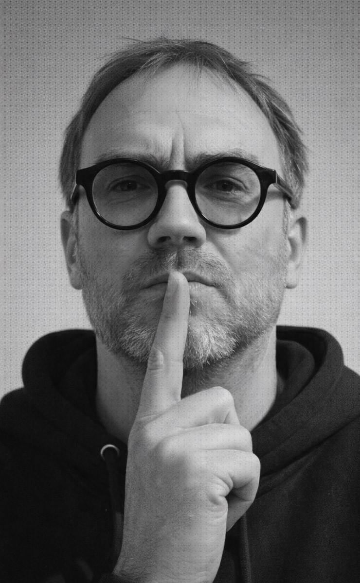 Man wearing glasses making shushing gesture in black and white portrait.