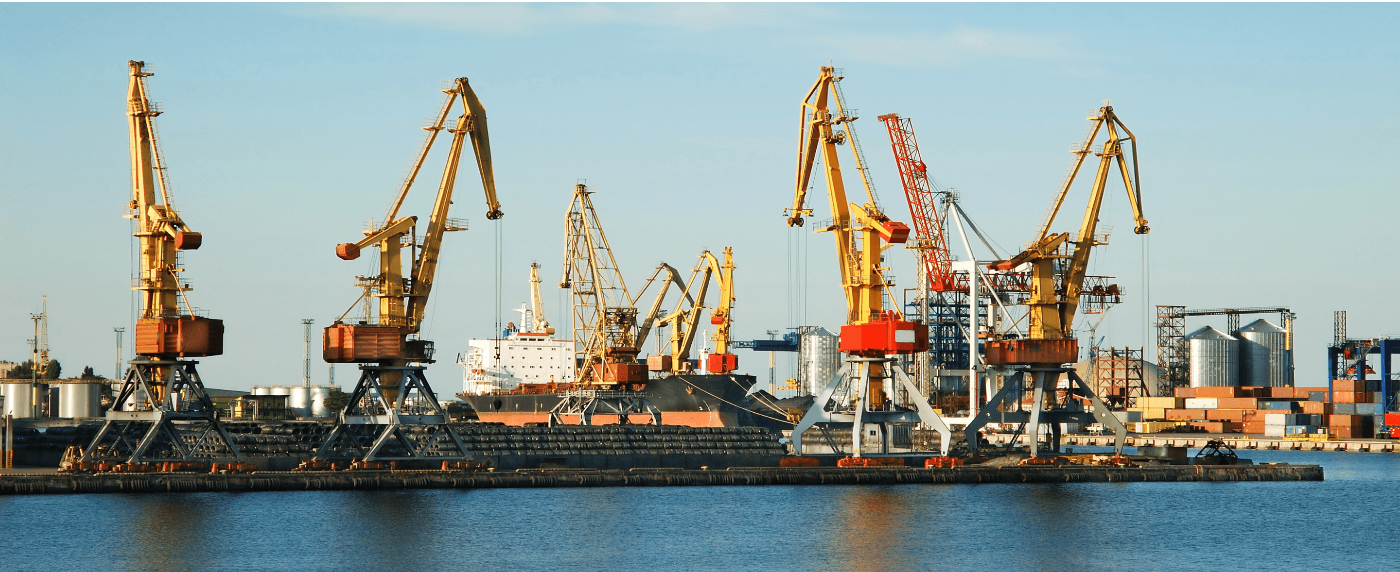 A harbor scene featuring multiple large cranes against a clear blue sky, with ships in the background.