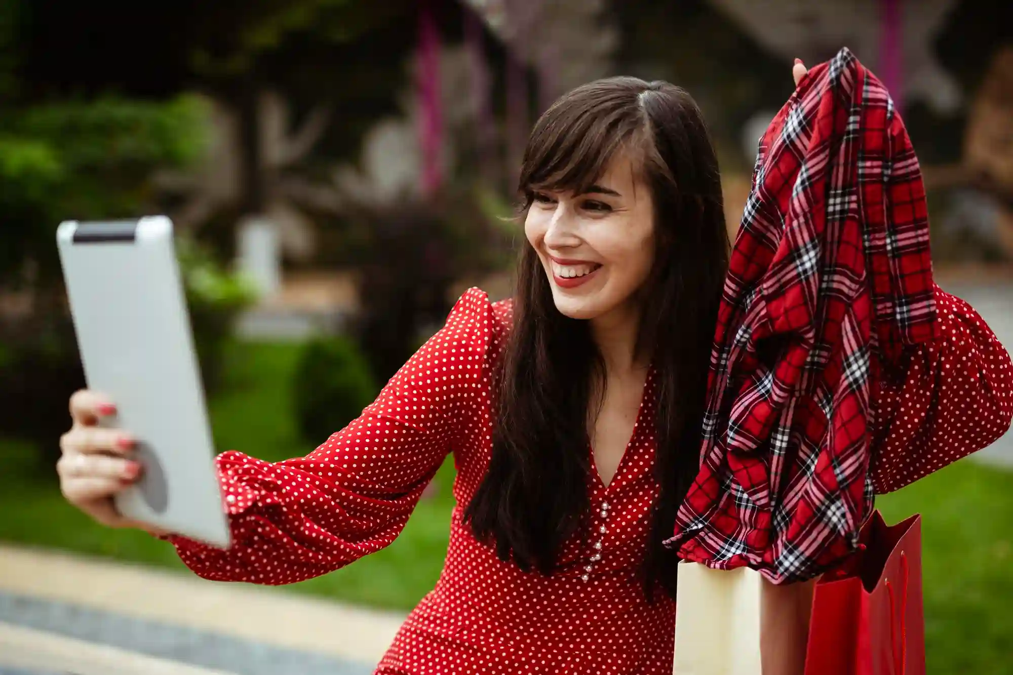 A smiling young woman in a red dress using a tablet to shop or video chat outdoors.